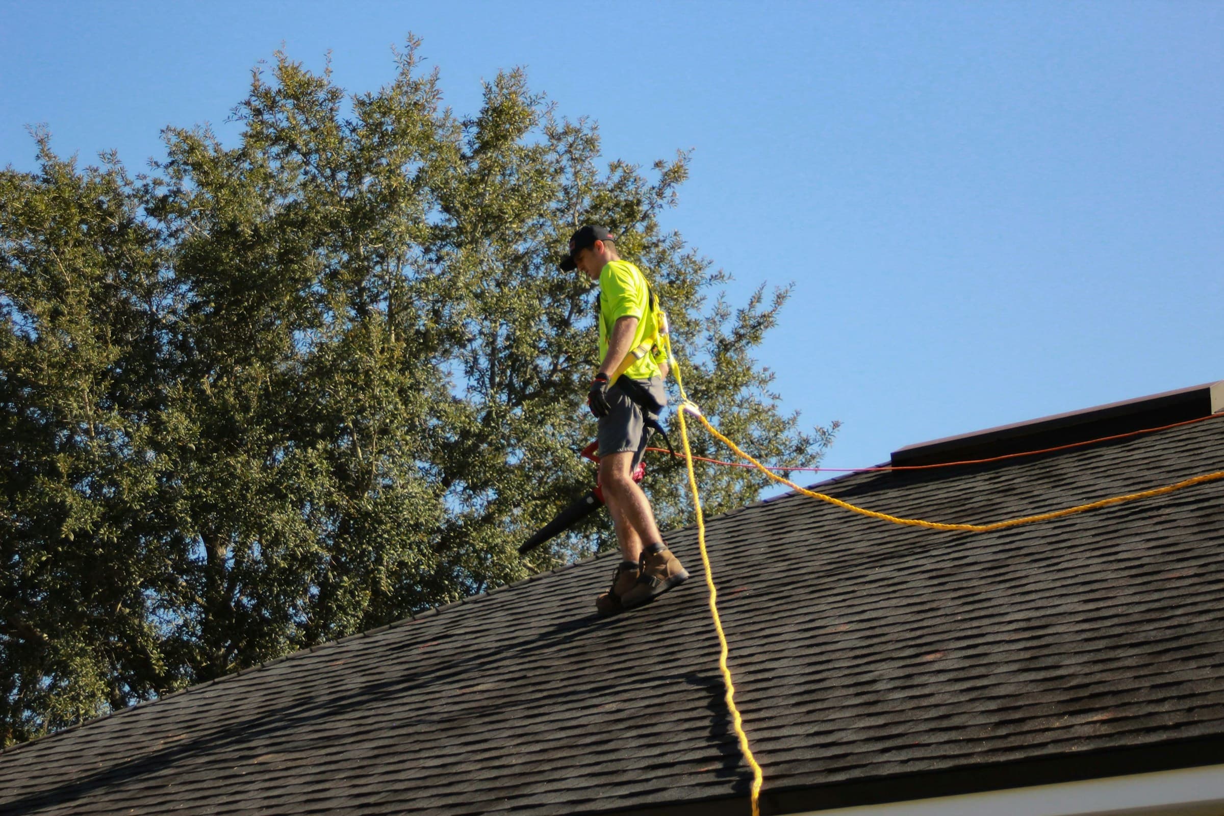 Roofer in a hi-vis shirt and safety harness working on a residential roof under a clear blue sky